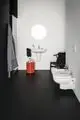 Waschtisch mit runder Waschschüssel und Armatur an der Wand, daneben ein roter Hocker und eine schwarze Stuhl. Badezimmer mit schwarzem Boden und hellen Wänden von Ideal Standard.